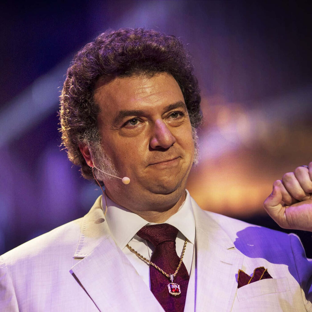 Danny McBride in sharp white suit deep burgundy tie gold accessories dramatic stage lighting wallpaper