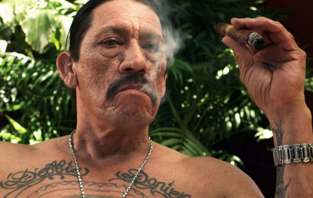 Danny Trejo smoking cigar with intense expression and tattoos against green foliage wallpaper