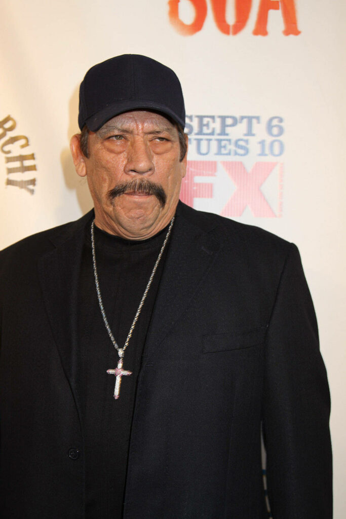 Danny Trejo intense look black jacket cap bold cross necklace rugged style wallpaper background