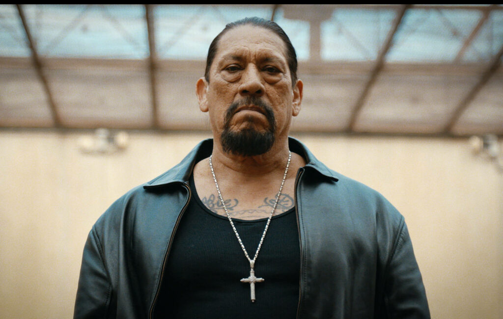 Danny Trejo fierce expression black leather jacket chain necklace urban sunlit background wallpaper