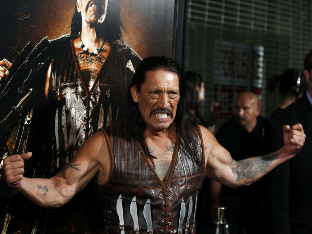 Danny Trejo flexes muscles wearing leather vest by Machete movie poster at gritty film event Wallpaper