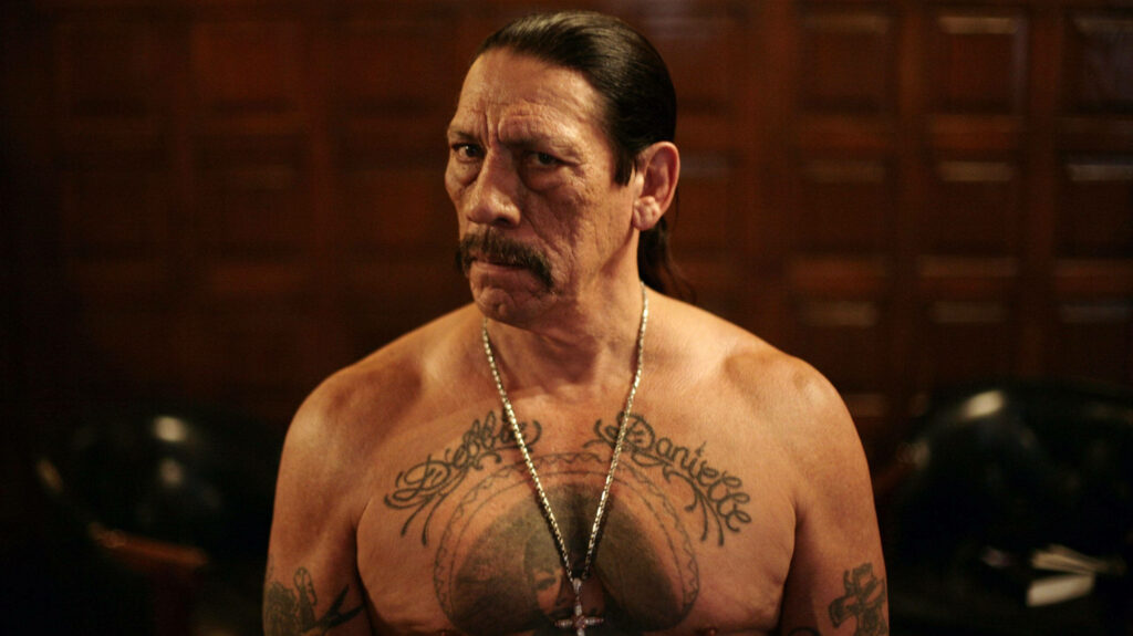 Danny Trejo topless showing muscular physique and bold tattoos against wooden wallpaper background