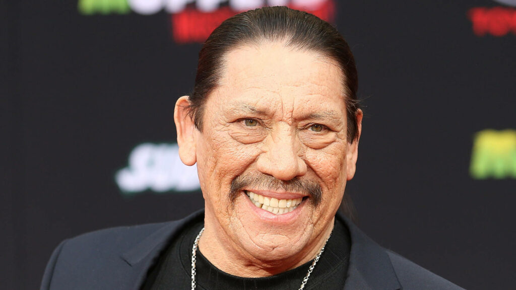 Danny Trejo smiling on red carpet vibrant wallpaper with colorful blurred event logos background