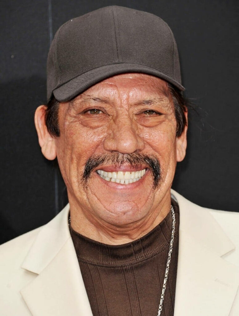 Danny Trejo joyful portrait wearing dark cap cream blazer vibrant wallpaper black background