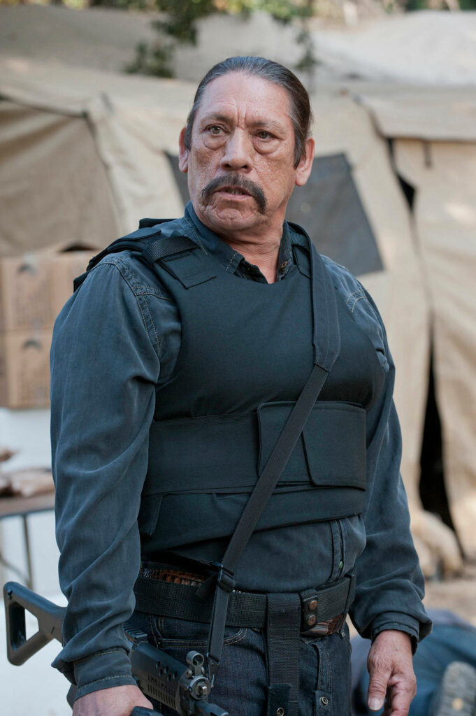 Danny Trejo wearing tactical vest and dark shirt in front of beige tent intense action star pose Wallpaper