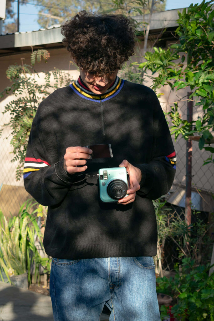 DannyLux outdoors holding vintage instant camera developing photo with natural sunlight and greenery Wallpaper