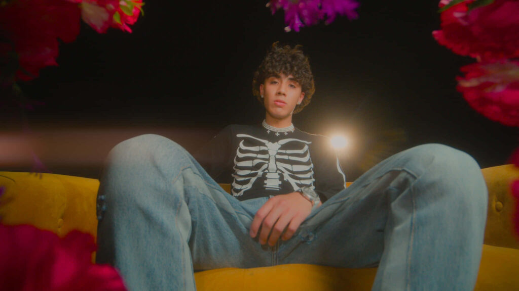 DannyLux sitting on yellow couch wearing skeleton shirt blue jeans colorful floral frame wallpaper