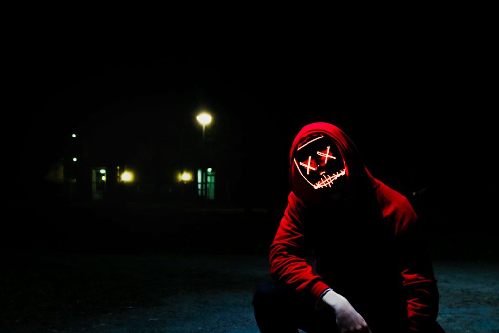 Figure in hoodie with glowing neon red X LED mask crouched in dark moody background wallpaper