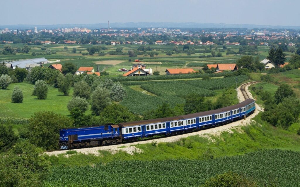 Aerial view of dark blue train curving through green fields near city with red roofs summer sky Wallpaper