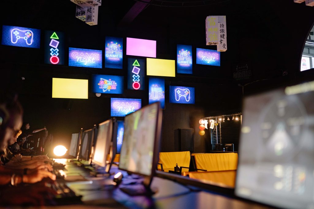 Dark neon gaming room with rows of monitors players and glowing Game Night controller wall panels Wallpaper