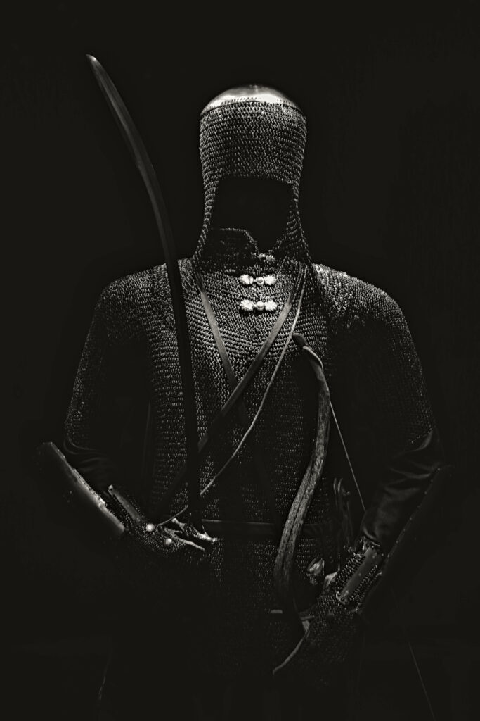 Mysterious warrior in black chainmail armor holding sword stands powerfully in dark shadows Wallpaper