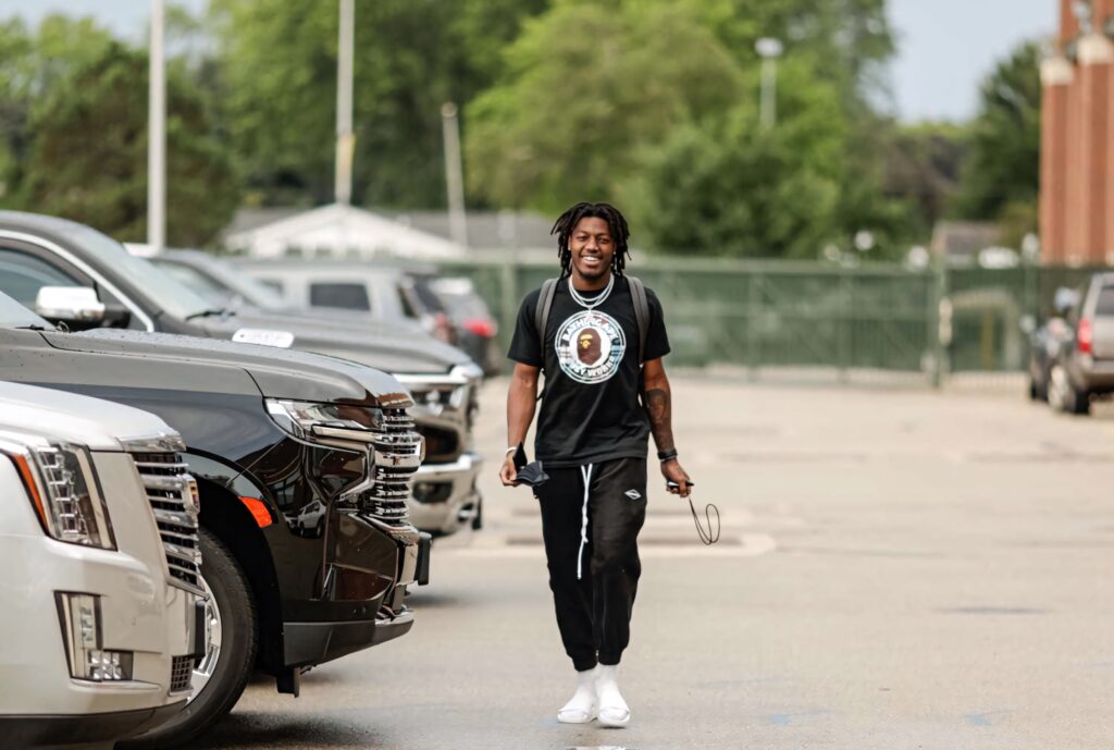 Darnell Savage Jr walking confidently in casual athletic wear near sleek cars and green trees Wallpaper