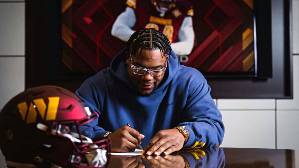 Daron Payne in blue hoodie signing contract with Washington Commanders helmet and photo background Wallpaper