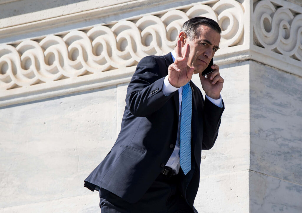 Darrell Issa in dark suit blue tie flashing peace sign talking on phone against white stone wall Wallpaper