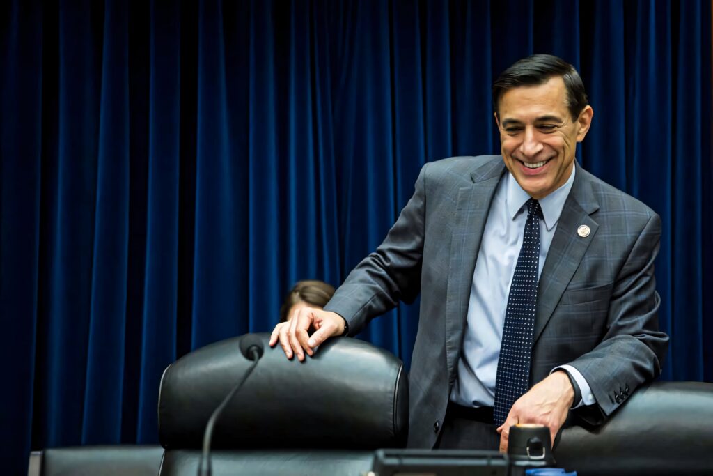 Darrell Issa smiling in formal attire standing before black chairs and deep blue curtain background Wallpaper