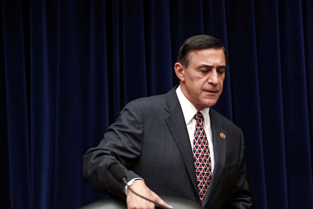 Darrell Issa in dark suit with patterned tie standing by chair blue curtains formal wallpaper