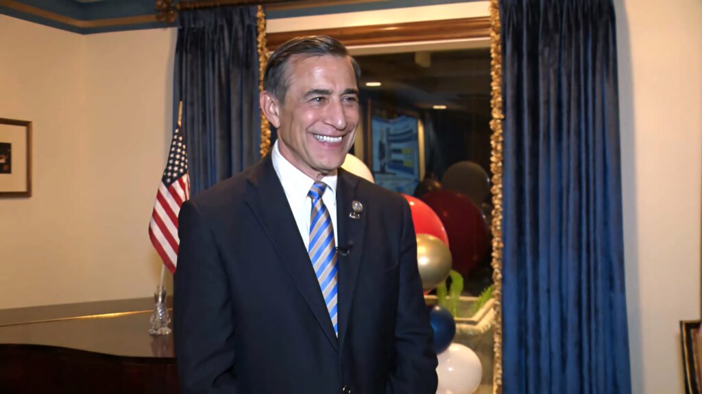 Darrell Issa in business suit delivering speech with US flag and blue curtains in formal room Wallpaper
