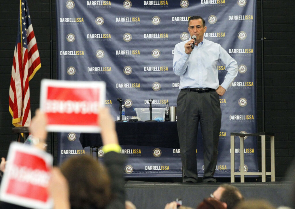 Darrell Issa speaking at political event with American flag and engaged crowd holding signs Wallpaper