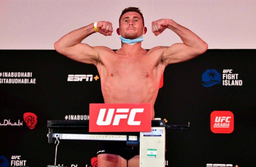 Darren Till flexing muscles at UFC weigh-in wearing blue face mask with intense fight atmosphere Wallpaper