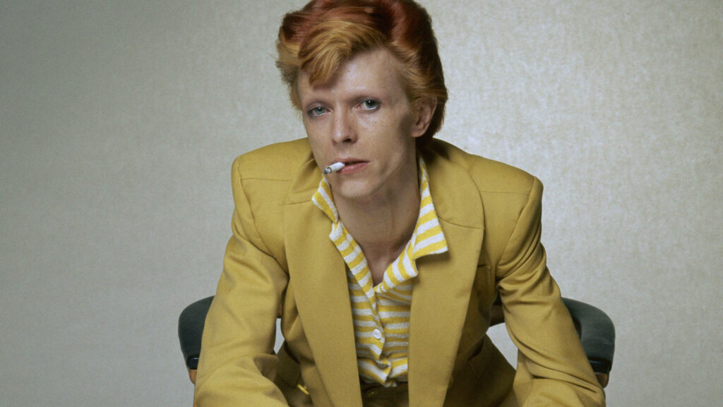David Bowie in bright yellow suit and striped shirt holding cigarette on neutral wallpaper background
