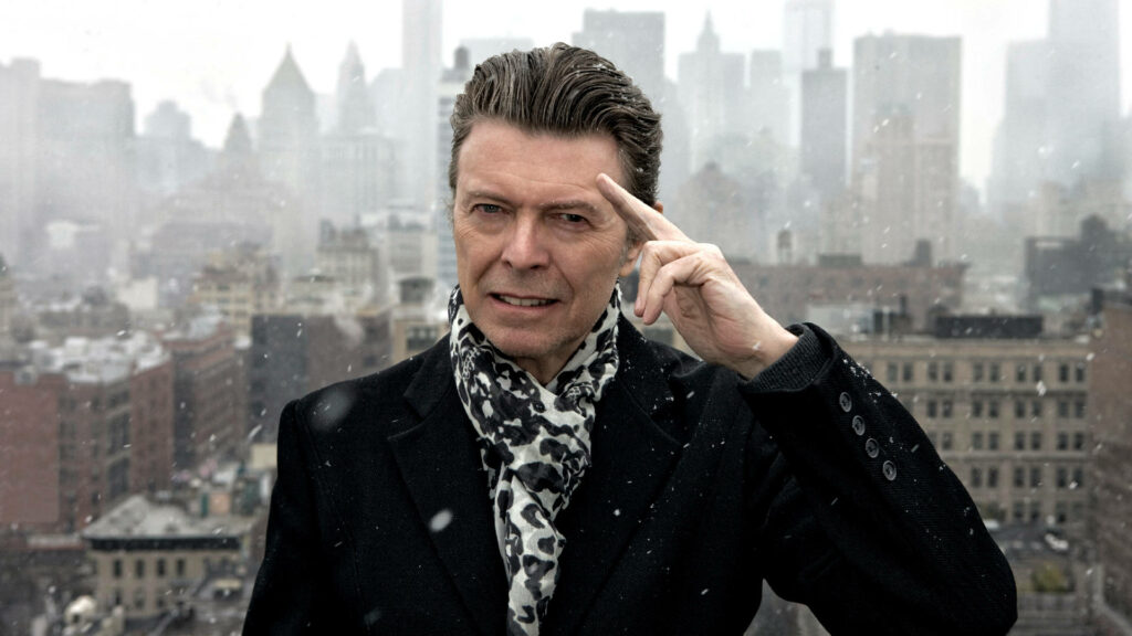 David Bowie on snowy rooftop wearing dark coat and patterned scarf with blurred city skyline Wallpaper