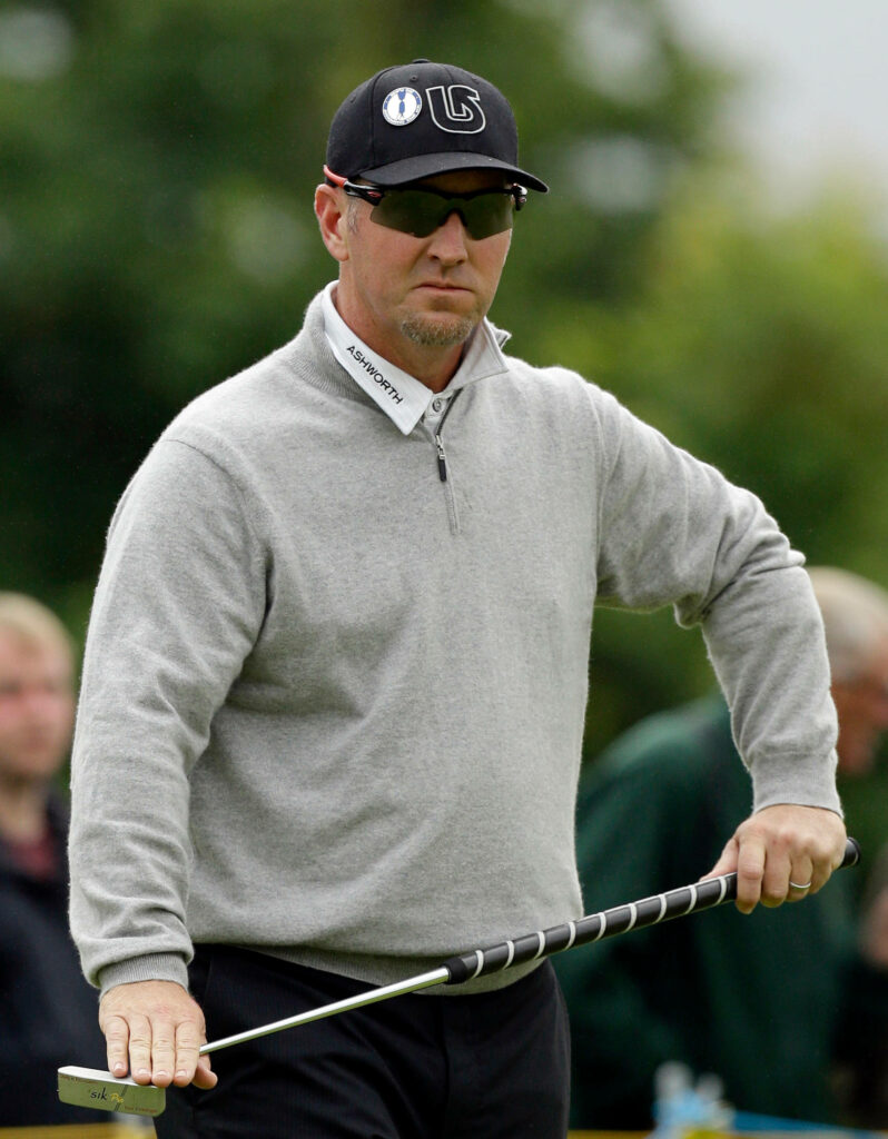 David Duval smirking in grey pullover and black cap holding golf club outdoors with green background Wallpaper