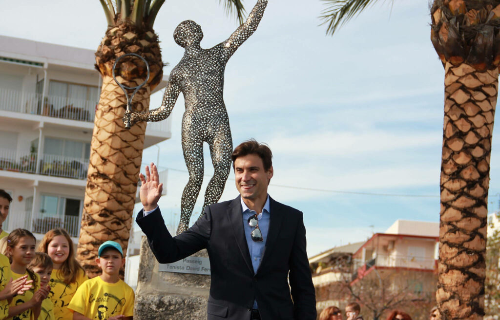 David Ferrer waving in dark suit by metallic tennis statue between palm trees at sunny event Wallpaper