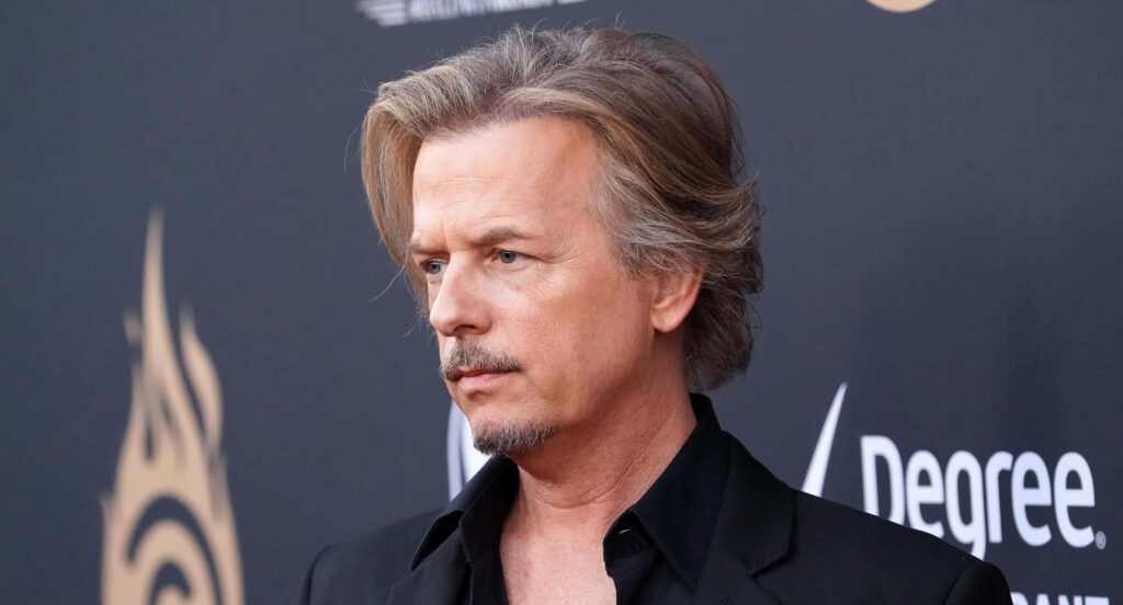 David Spade in black suit with thoughtful expression on dark stylish wallpaper background portrait