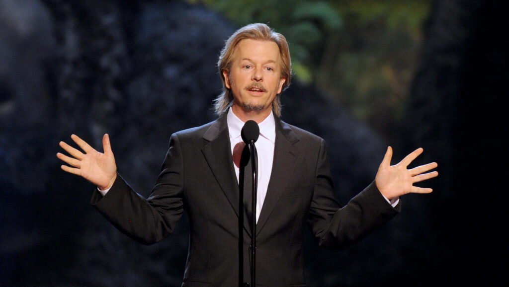 David Spade in black suit delivering comedic performance at microphone with blurred nature backdrop Wallpaper