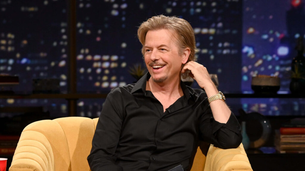 David Spade smiling on yellow sofa wearing black shirt gold watch cityscape background talk show Wallpaper