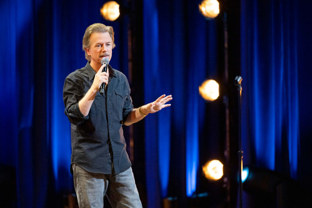 David Spade performing stand-up comedy on stage with blue curtain and warm stage lights Wallpaper