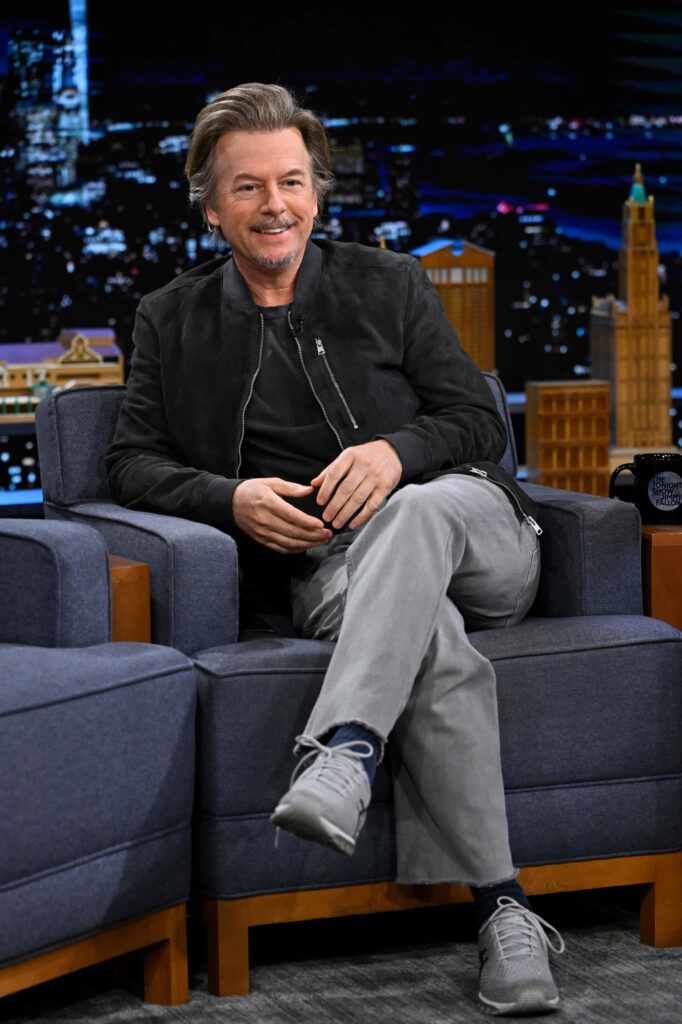 David Spade relaxed on talk show set wearing black jacket grey pants vibrant cityscape background Wallpaper