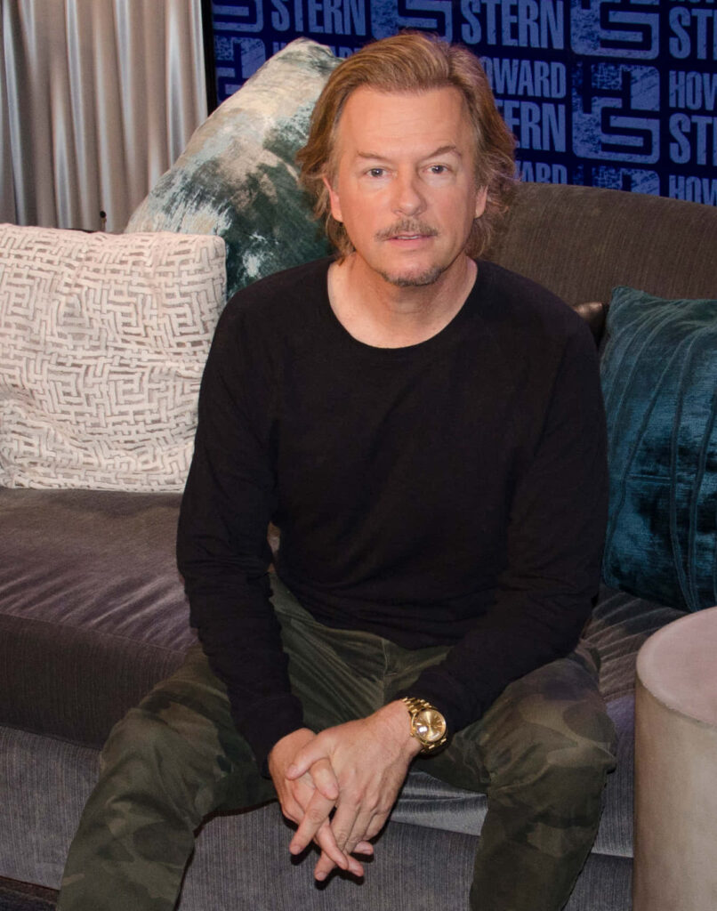 David Spade seated on modern sofa with camo pants black shirt blue Howard Stern sign background Wallpaper
