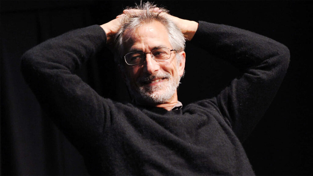 David Strathairn smiling warmly in black sweater relaxed pose against dark wallpaper background