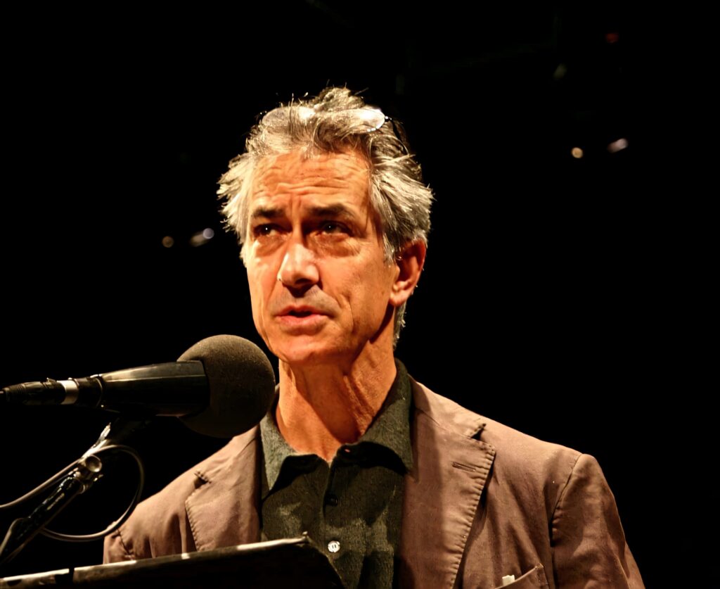 David Strathairn at microphone in dark blazer dramatic lighting cinematic wallpaper background