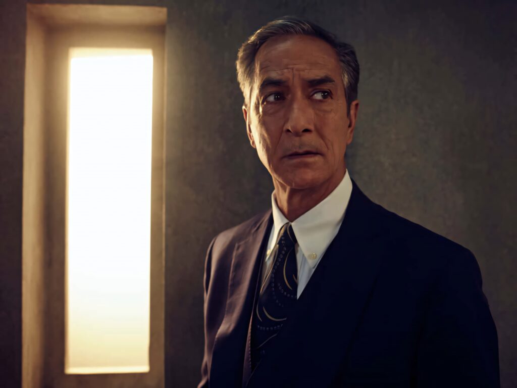 David Strathairn in sharp suit in dim room with warm wallpaper illuminated by soft window light