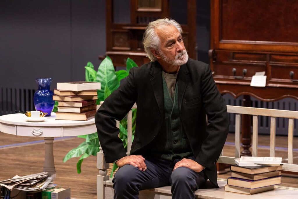 David Strathairn seated on wooden bench in warm book-filled room with rustic furniture and plants Wallpaper