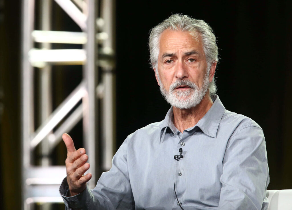 David Strathairn in light gray shirt against soft lit metal frame backdrop thoughtful expression Wallpaper
