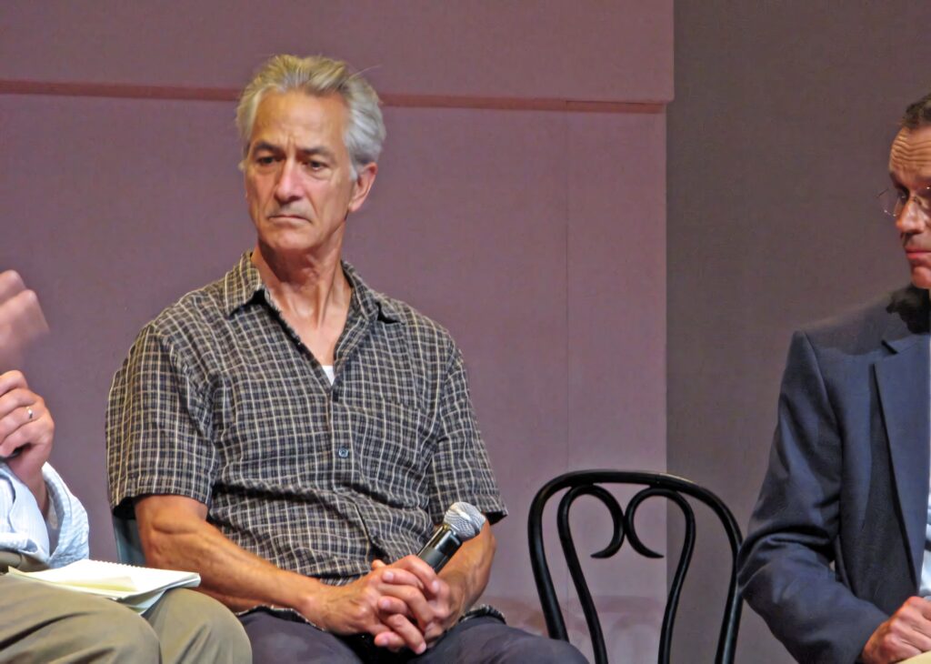 David Strathairn in checkered shirt speaking on stage during thoughtful panel discussion wallpaper
