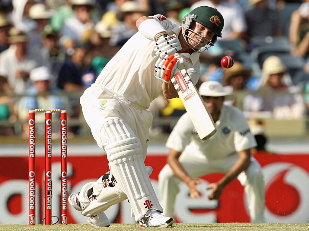 David Warner in Australian Test uniform mid-swing hitting cricket ball with intense focus outdoors Wallpaper