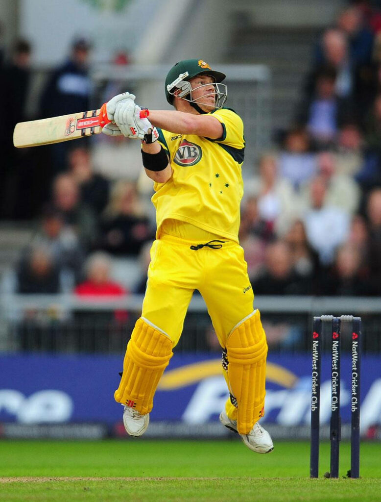 David Warner mid-leap batting in yellow Australia cricket kit with vibrant stadium background Wallpaper
