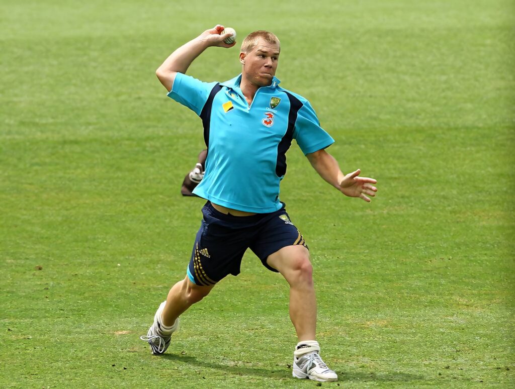 David Warner in Australian cricket training kit mid-action on vibrant green field with natural light Wallpaper