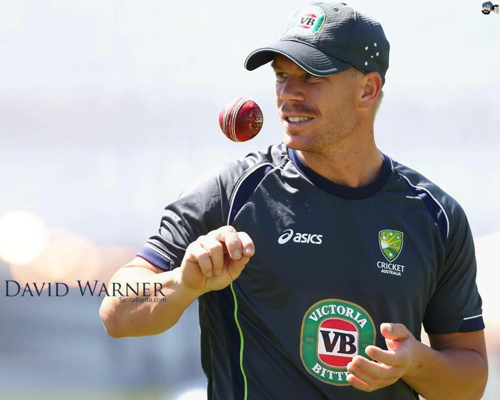 David Warner in Australian cricket training kit tossing red ball mid-action with sunlit background Wallpaper