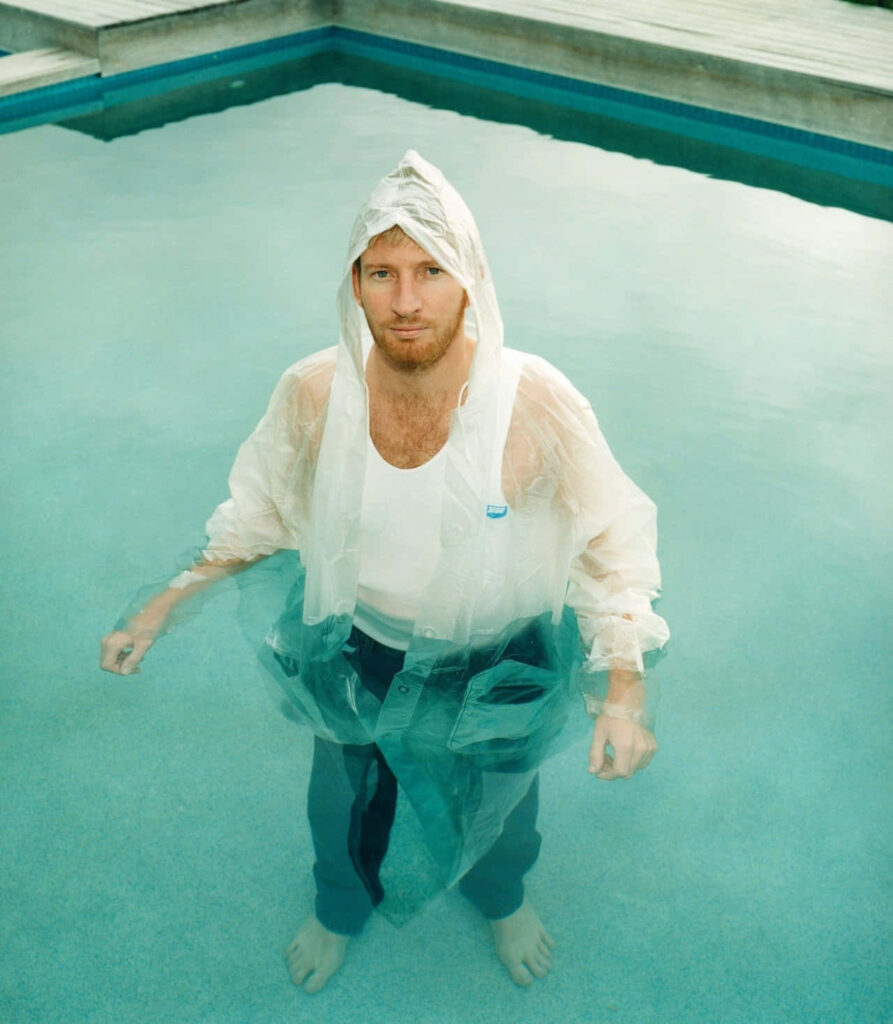 David Wenham barefoot in pool wearing translucent raincoat casual clothes calm blue water serene outdoors Wallpaper