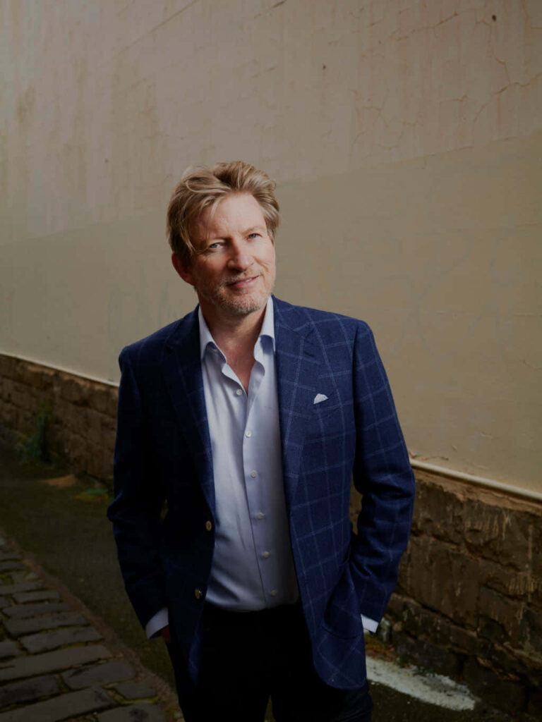 David Wenham in blue checkered suit jacket against rustic wall natural light portrait wallpaper