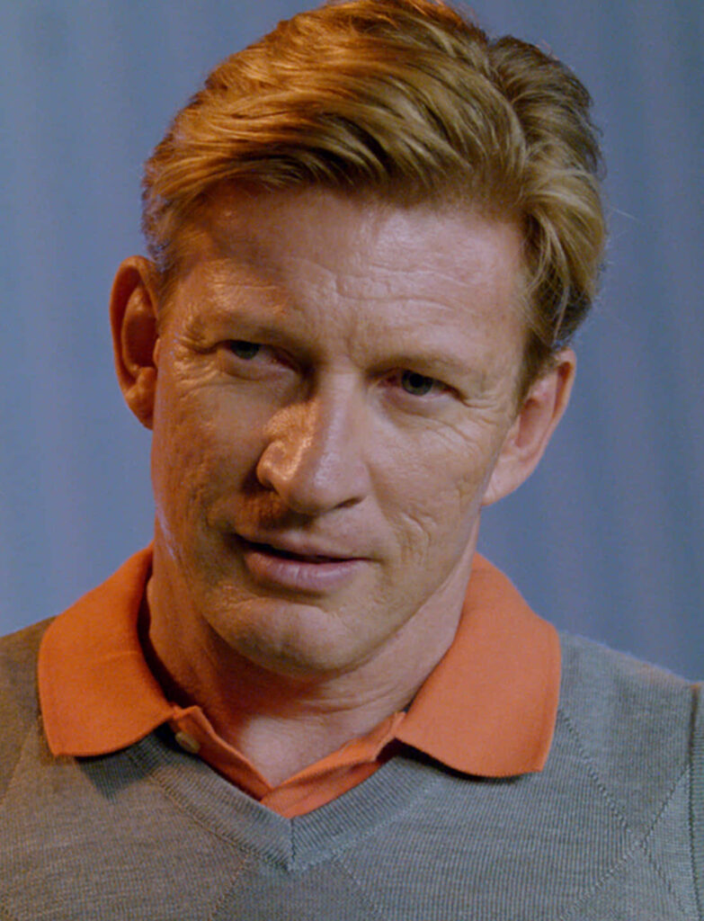 Close-up portrait of David Wenham in casual orange shirt and gray sweater on soft blue background Wallpaper