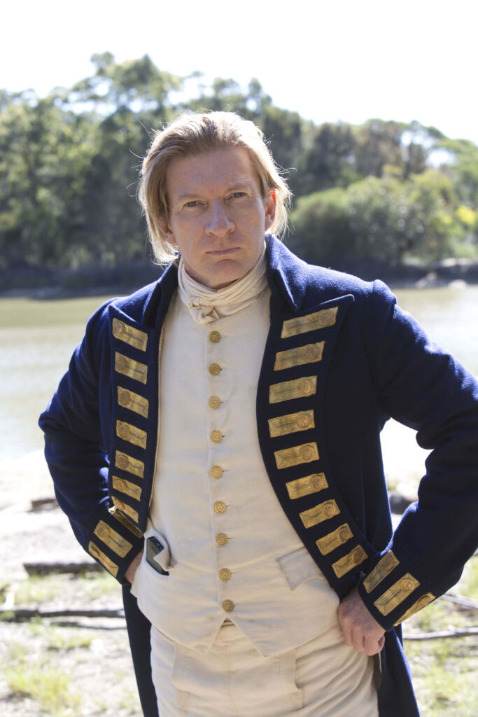 David Wenham in historical navy blue military coat with gold accents by riverside trees daylight Wallpaper