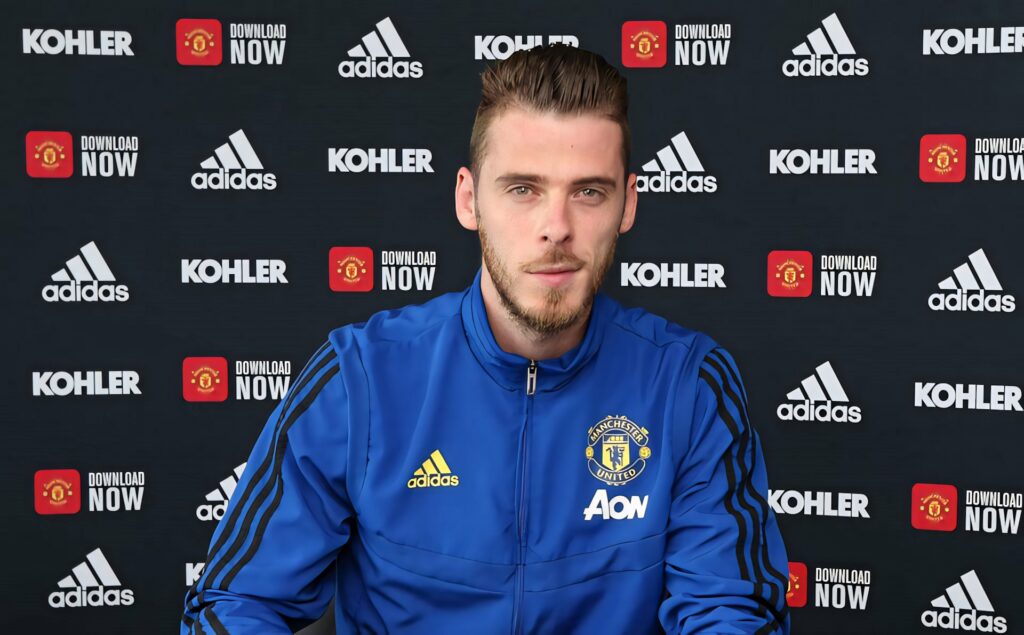David de Gea in blue Manchester United jacket at press conference with branded backdrop wallpaper