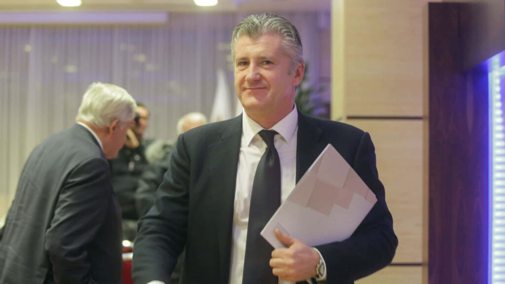 Davor Šuker in dark suit smiling confident at formal event holding documents modern indoor setting Wallpaper