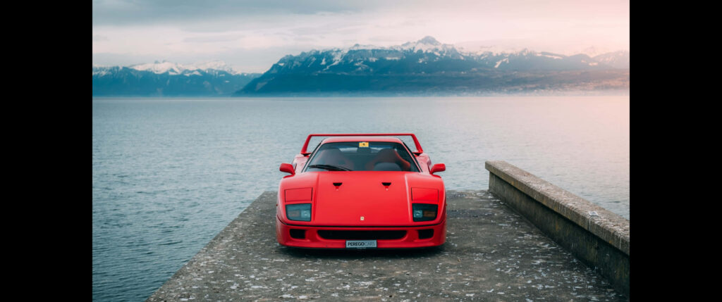 Cruising Paradise: Red Ferrari F40 Speeding Through Scenic Waterscape with Majestic Mountain Backdrop Wallpaper